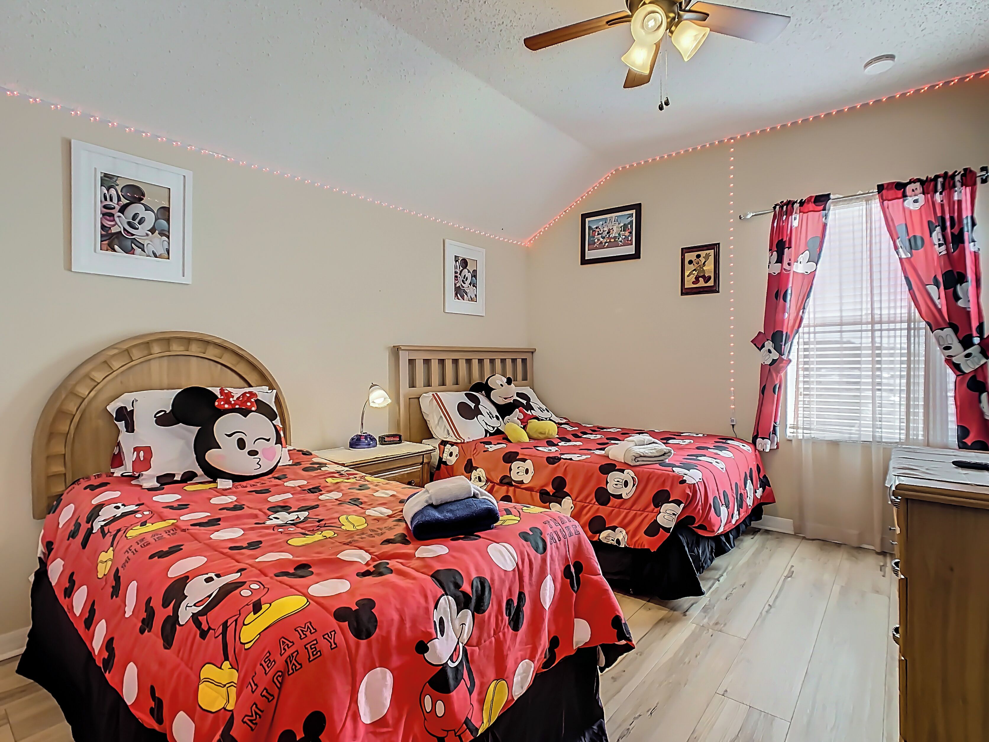 Una habitación con dos camas, ambas cubiertas con ropa de cama temática de Mickey Mouse, un ventilador de techo y luces colgantes.