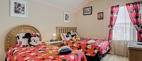 Una habitación con dos camas, ambas cubiertas con ropa de cama temática de Mickey Mouse, un ventilador de techo y luces colgantes.