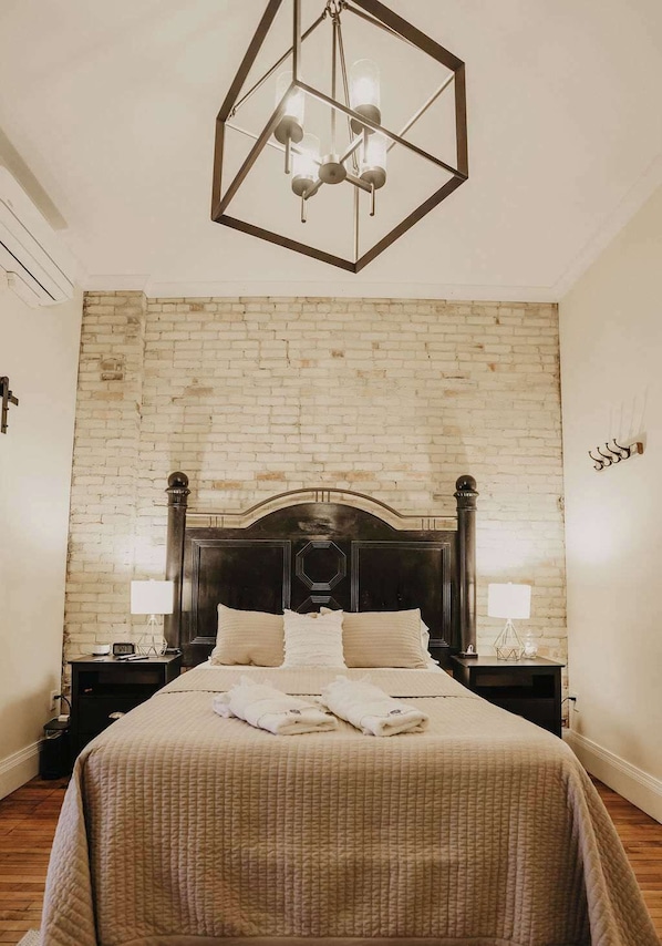 Un dormitorio con pared de ladrillo, cabecera de madera, cama con colcha, dos mesitas de noche con lámparas y un aplique de techo geométrico.