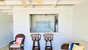 Un salon moderne avec un canapé blanc, deux fauteuils en bois, une petite table basse et un coin cuisine doté d’un plan de travail blanc et de décorations murales aux motifs géométriques.