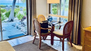 Une pièce avec un ordinateur portable posé sur une table, des chaises en osier, une lampe et une vue sur la plage et les palmiers, visible à travers de grandes fenêtres.