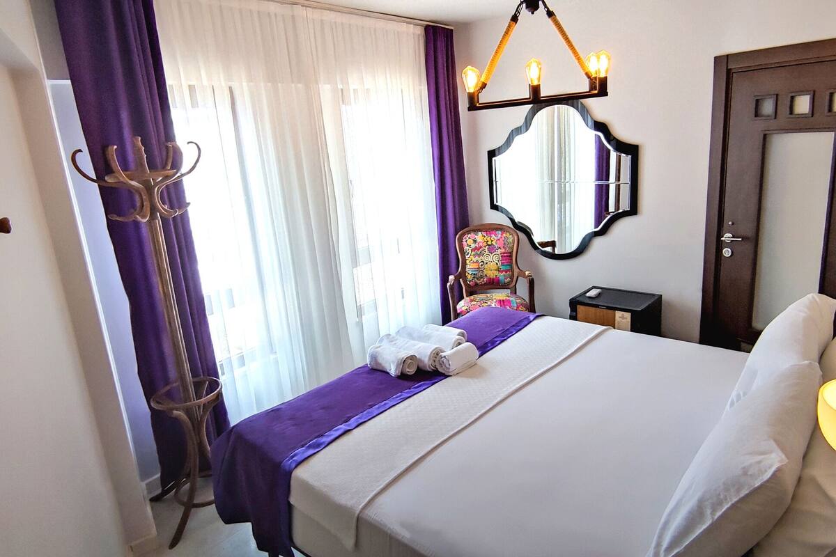 A hotel room with a bed, a chair, a mirror, a door, and a lamp.