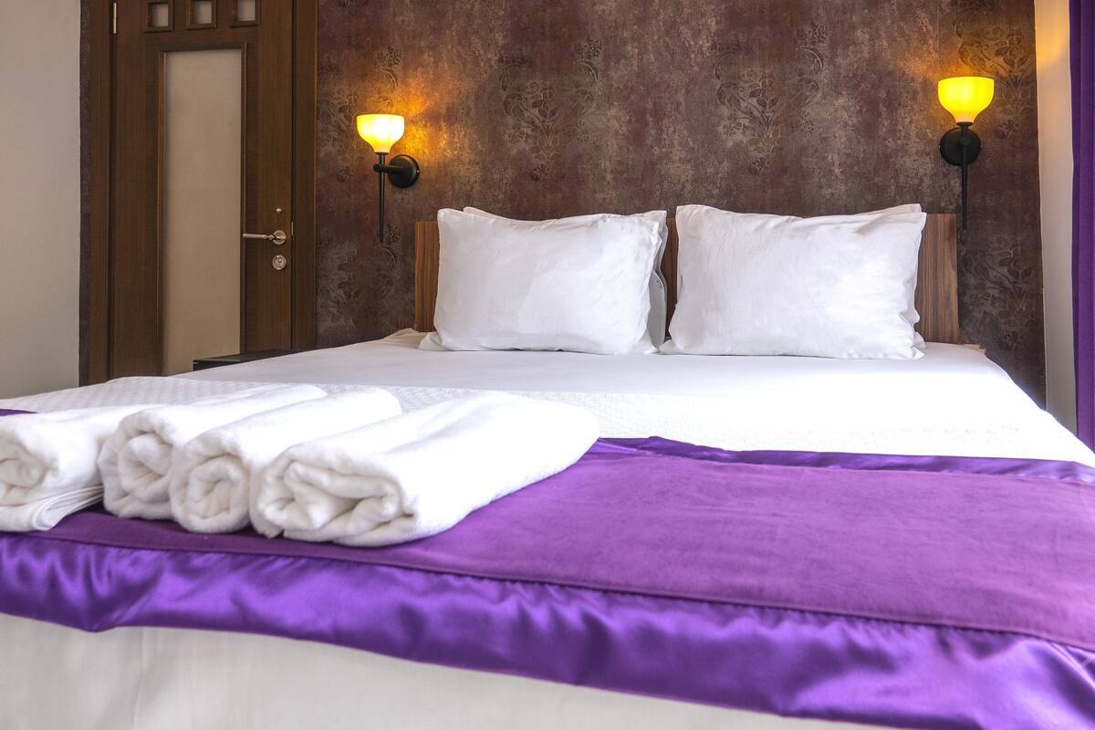 A neatly made bed with white linens, a purple bedspread, and rolled towels on the side.
