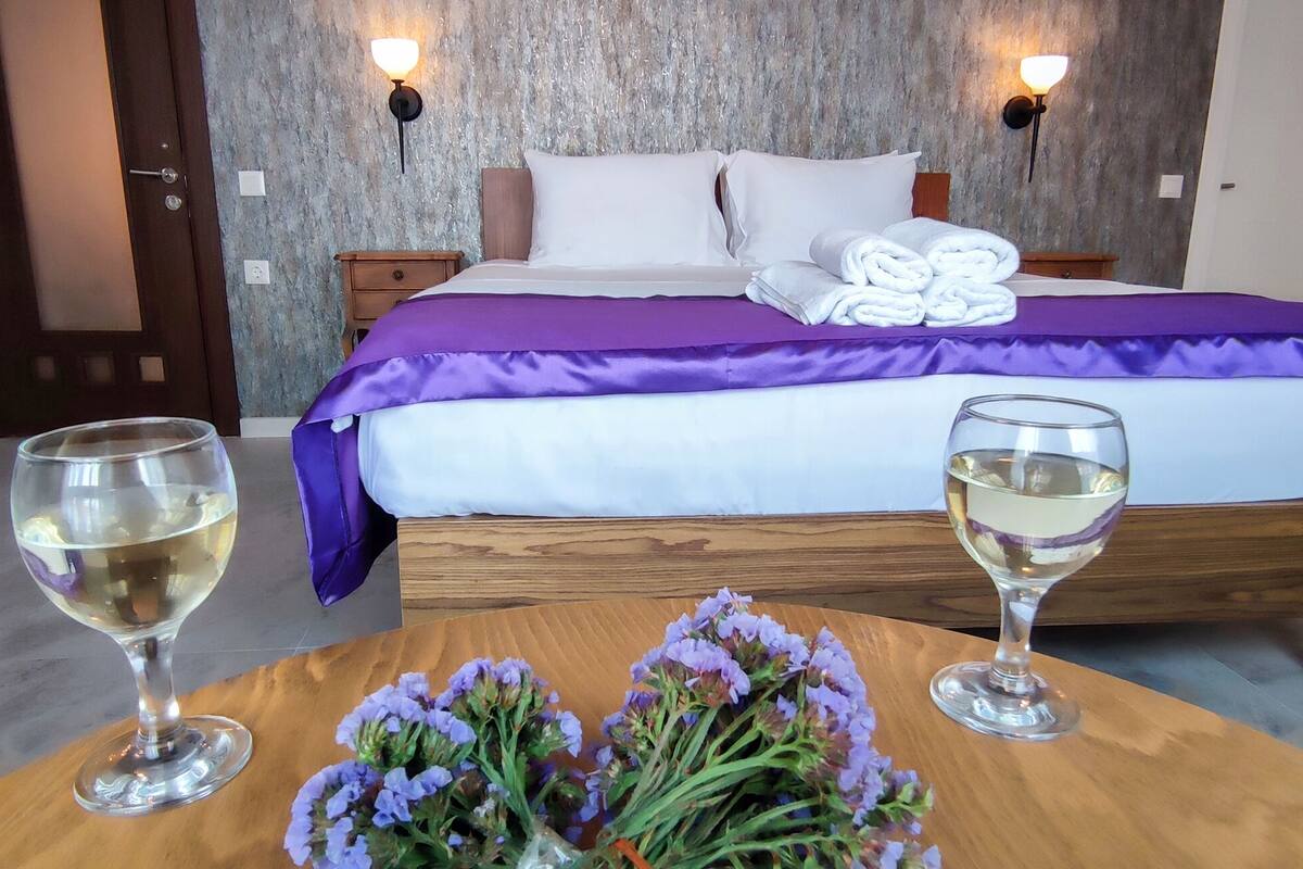 A hotel room with a bed, two bedside lamps, a wooden nightstand, a table with two glasses of white wine, and a bouquet of purple flowers.