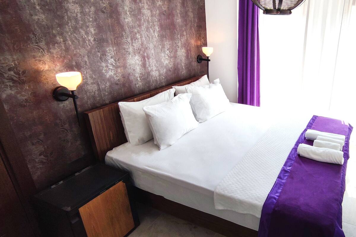 A neatly made bed with white linens and a purple bedspread, a nightstand with a lamp, and a window with purple curtains.