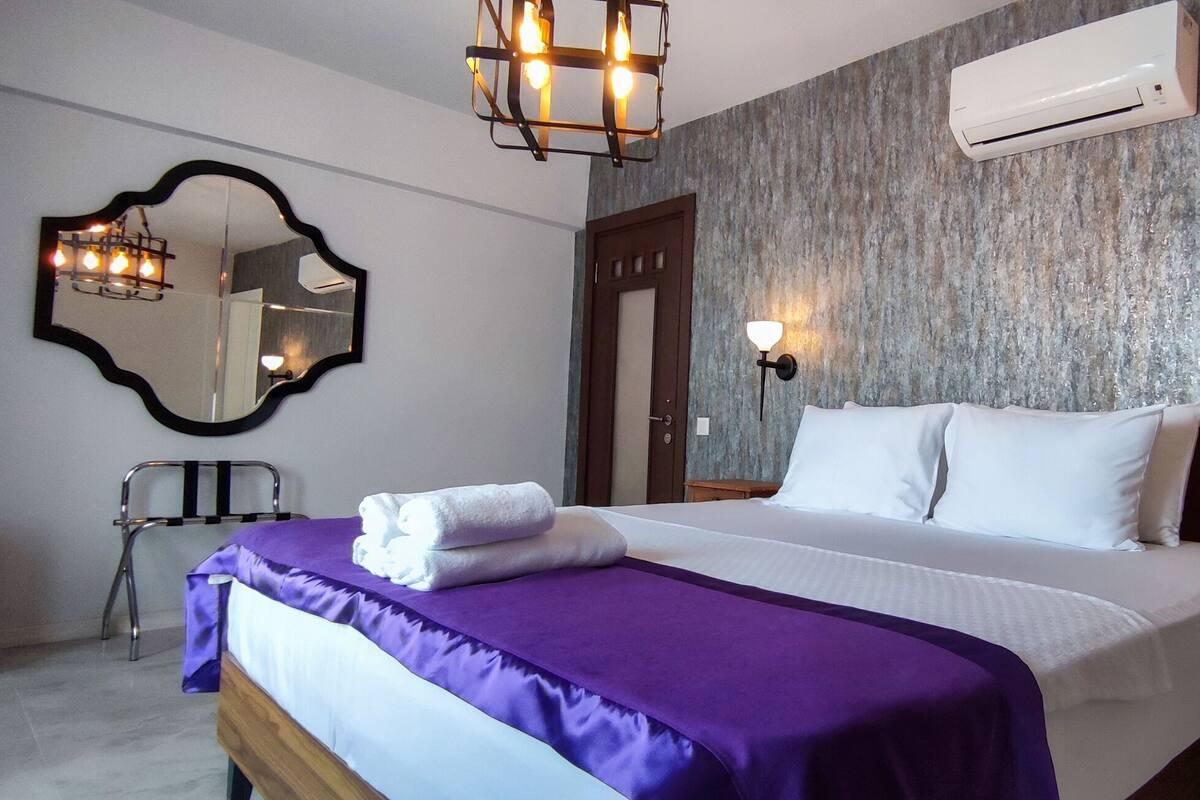 A hotel room with a bed, a mirror, a chair, an air conditioner, and wall-mounted lights.