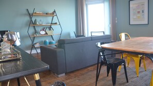 A modern living room with a wooden dining table, a grey sofa, and a black and yellow chair. There is a bookshelf with various items and a window with curtains.