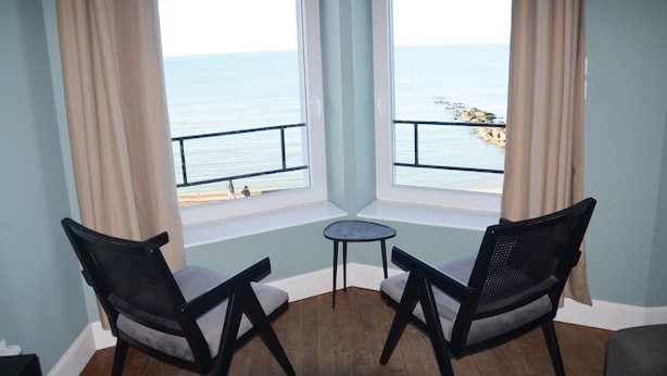 A room with a view of the sea, two chairs, a small table, and a window with curtains.