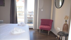 A hotel room with a bed, a red armchair, a round table with a lamp, and a view of buildings through the window.