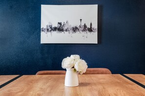 Un vase blanc orné de fleurs, posé sur une table en bois, devant un mur bleu sur lequel est accroché un tableau représentant un paysage urbain.