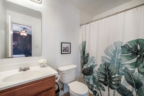 Une salle de bain avec un lavabo blanc, des toilettes, un tableau encadré au mur et un rideau de douche orné d’un motif de feuilles.