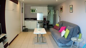 Un espacio habitable compacto con kitchenette, una mesa de comedor, un sofá con cojines coloridos y una pequeña mesita auxiliar.