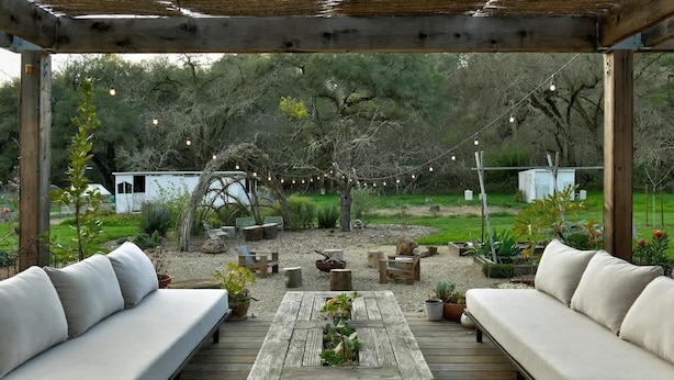 A covered outdoor seating area with two sofas and a wooden table, surrounded by plants and a rustic setting. - Healdsburg Homestead with Pool, Hot Tub and Tastings Included (Healdsburg)
