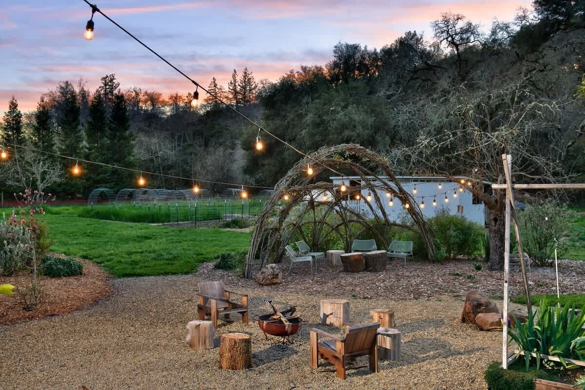 An outdoor seating area with wooden chairs and a fire pit, surrounded by a natural landscape and string lights.
