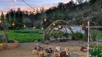 An outdoor seating area with wooden chairs and a fire pit, surrounded by a natural landscape and string lights.