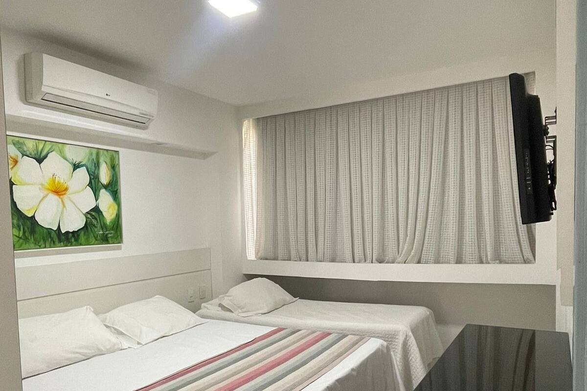 A hotel room with two beds, a painting of flowers, an air conditioning unit, a television, and a window with curtains.
