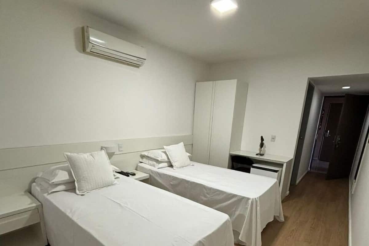 A hotel room with two single beds, a desk, a chair, a wardrobe, and an air conditioning unit.