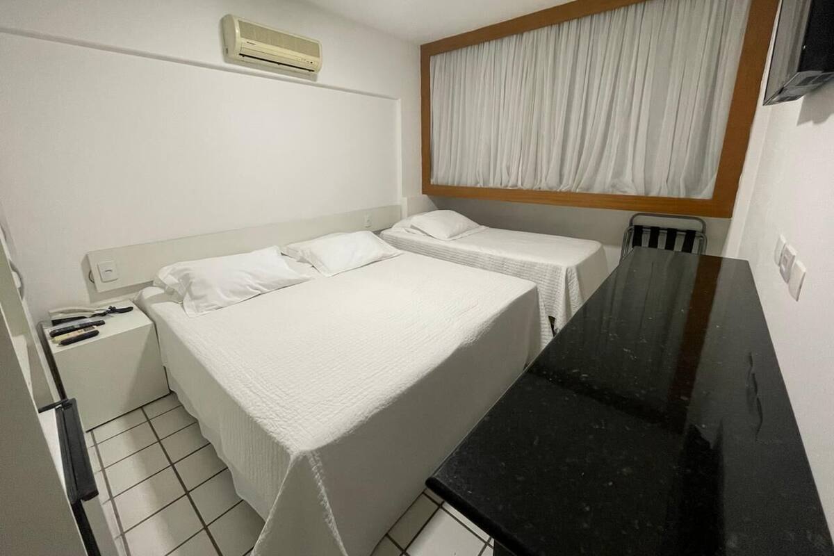 A small, single-bed room with a white bed, a black countertop, and a window with white curtains.