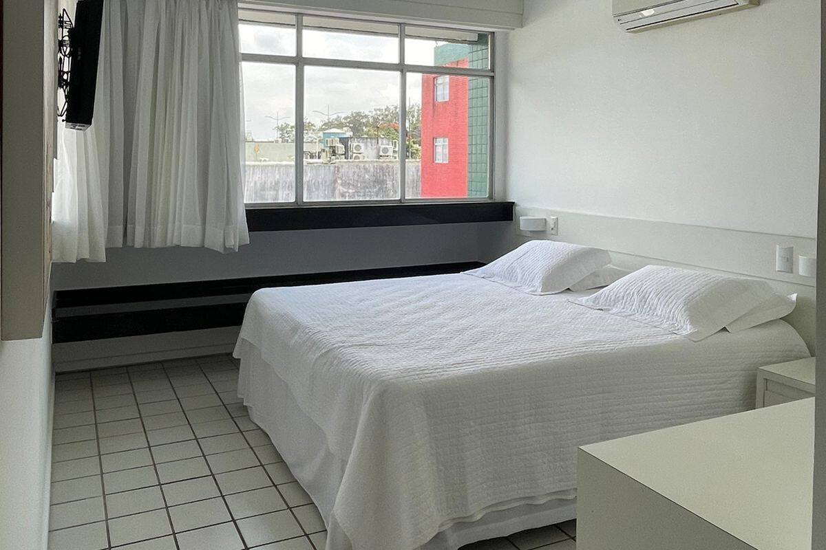 A hotel room with a bed, a window with curtains, a wall-mounted air conditioner, and a tiled floor.