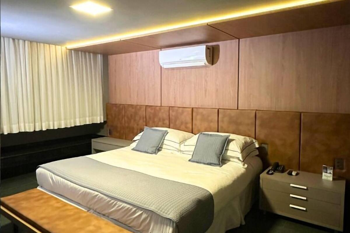 A hotel room with a large bed, bedside tables, a bench, and an air conditioning unit.