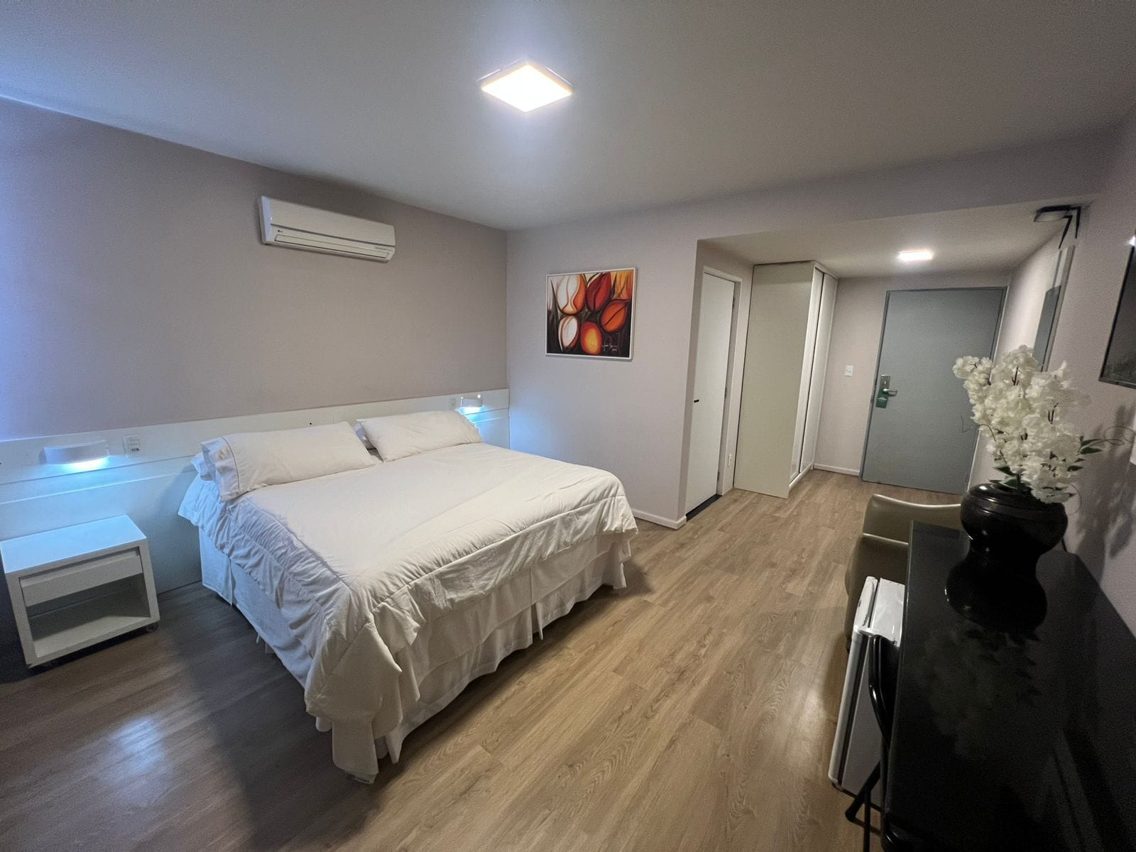 Un dormitorio con cama, mesitas de noche, un cuadro, una puerta y un florero con flores.