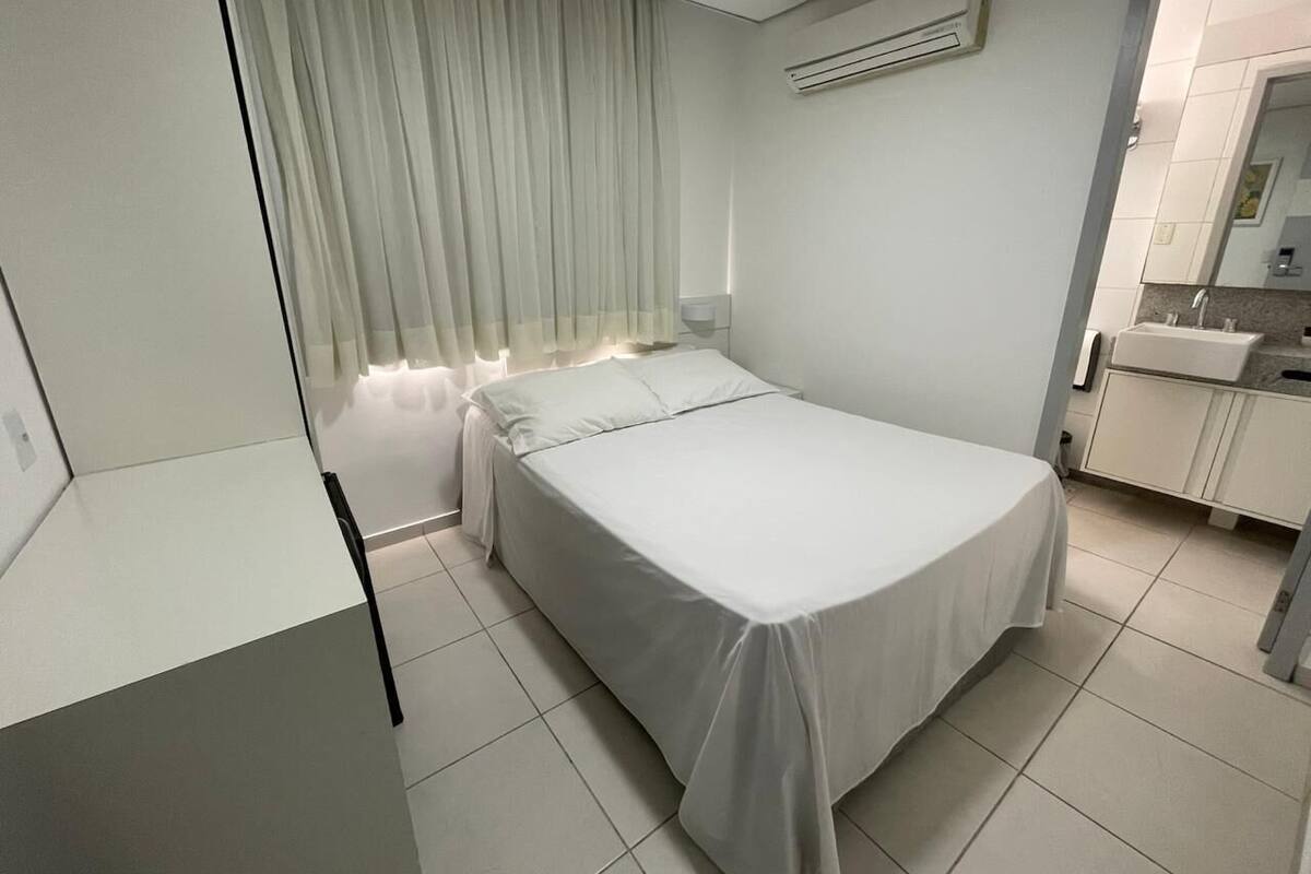 A modern hotel room with a neatly made bed, a bedside table, a window with sheer curtains, a wall-mounted air conditioner, and a bathroom with a sink and mirror.