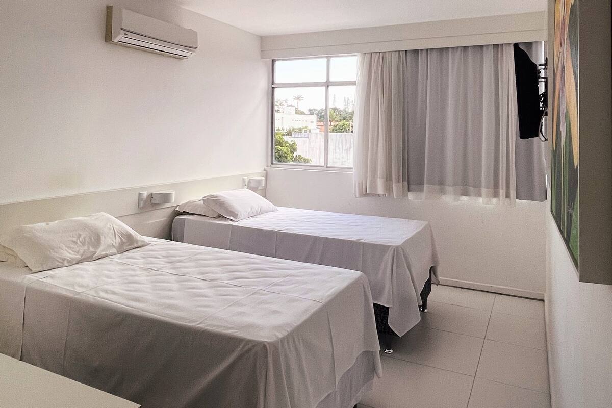 A hotel room with two single beds, a window with curtains, a wall-mounted air conditioner, and a flat-screen TV.