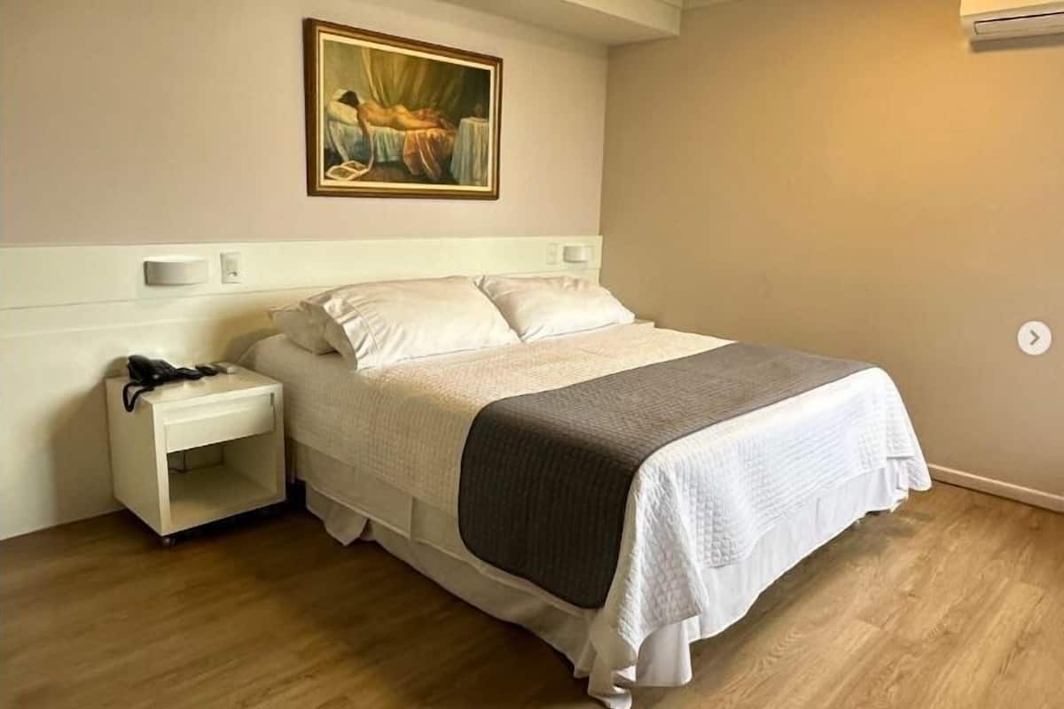 A bedroom with a bed, a nightstand, a painting, and an air conditioning unit.