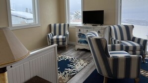 Una sala de estar acogedora con sillones a rayas, un televisor sobre una cómoda y ventanas grandes con vistas a árboles cubiertos de nieve.