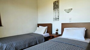 Habitación de hotel con dos camas individuales, cada una con una colcha gris acolchada y almohadas blancas. Hay una mesita de noche con una lámpara entre las camas, y una lámpara de pared encima de la mesita.