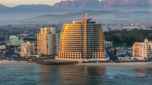 A modern hotel with a unique curved architecture, situated by a waterfront with a view of distant mountains. - Beachfront Luxury 1,300 Sq Ft, Workspace, Air Con & False Bay Views (Cape Town)