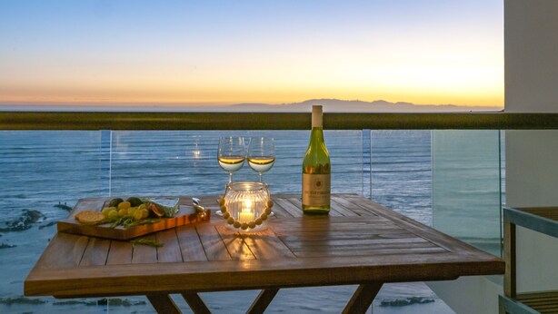 A wooden table with a bottle of wine, two glasses, a candle, and a fruit platter by a seaside view. - Beachfront Luxury 1,300 Sq Ft, Workspace, Air Con & False Bay Views (Cape Town)