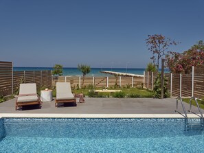 Un espace piscine comprenant deux chaises longues et une petite table, avec vue sur une plage offrant une vue dégagée sur la mer et une promenade en bois.