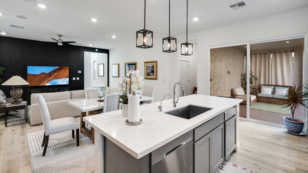 Una cocina moderna con una isla central, encimeras blancas y electrodomésticos de acero inoxidable. Hay un comedor con una mesa y sillas blancas, y una sala de estar con sofá y televisor.