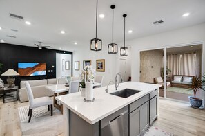 Een moderne keuken met een centraal eiland, witte aanrechtbladen en roestvrijstalen apparatuur. Er is een eethoek met een witte tafel en stoelen, en een woonkamer met een bank en een televisie.
