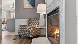 Una sala de estar con chimenea, un sillón estampado, una mesita de noche con lámpara y un cuadro en la pared.