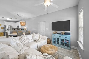 A modern living room with a white sofa, a dining area, a kitchen, and a TV mounted on the wall.
