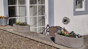 Un bâtiment à l’extérieur blanc, doté de grandes fenêtres et d’une porte ornée d’une grille métallique. On y trouve deux jardinières en béton remplies de fleurs et une chaise en osier à l’extérieur.
