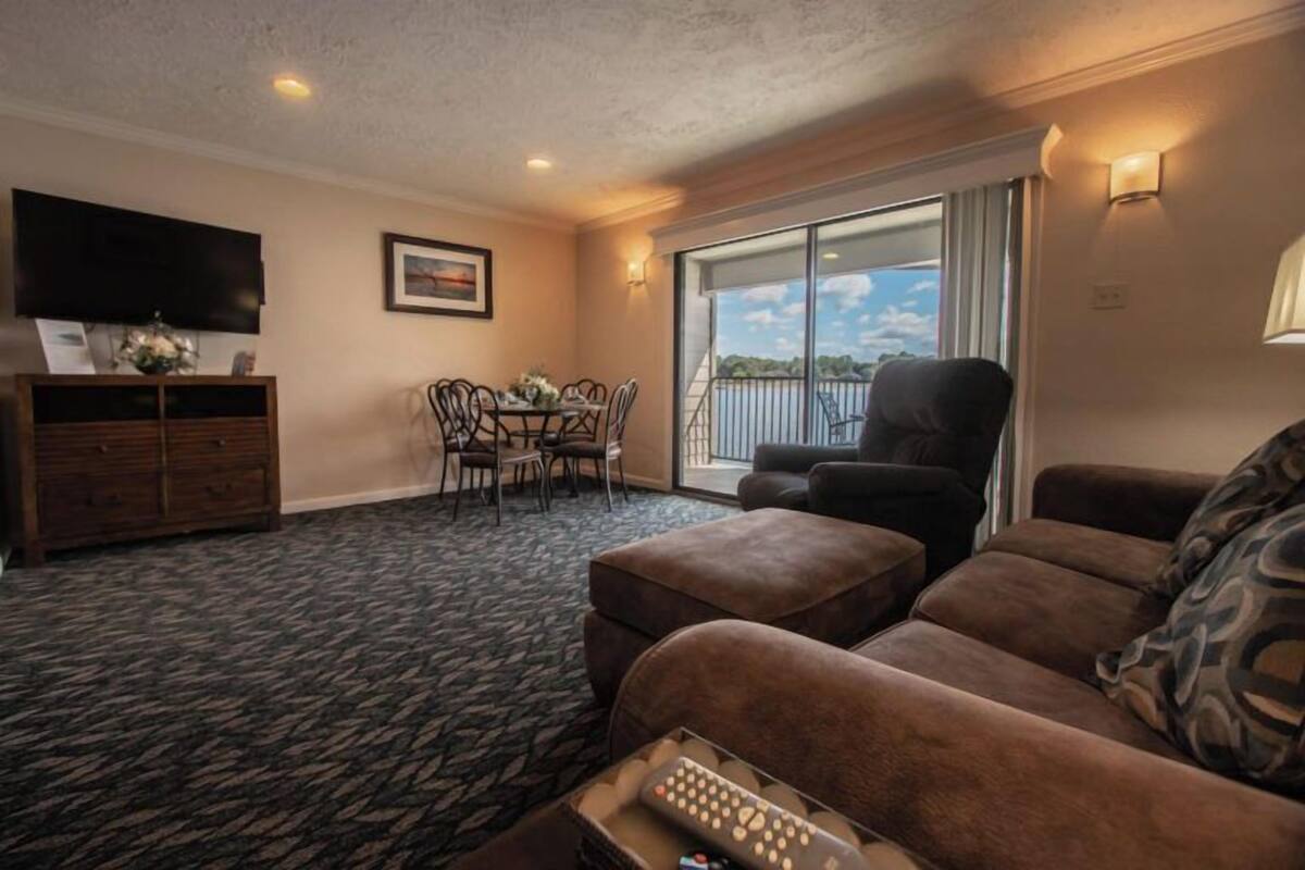 A living room with a sofa, ottoman, and a television on a stand. There is a dining table with chairs and a window with a view of the outdoors.