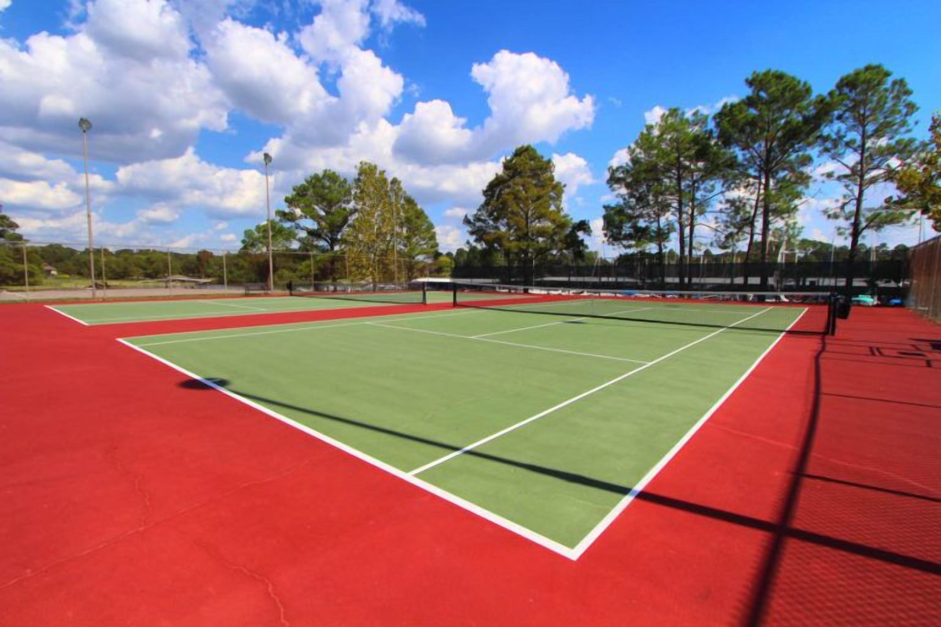 A tennis court with a red surface and green boundary lines, surrounded by trees and a clear sky.