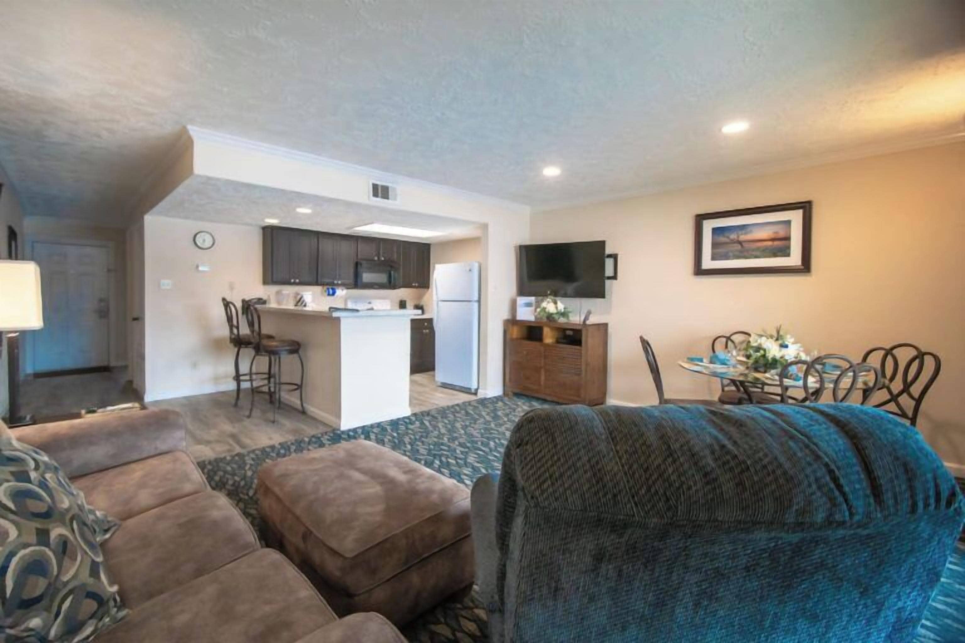 A living room with a sofa, ottoman, and a blue chair. There is a kitchen area with a refrigerator, microwave, and a dining table with chairs. A TV is mounted on the wall above the dining table.