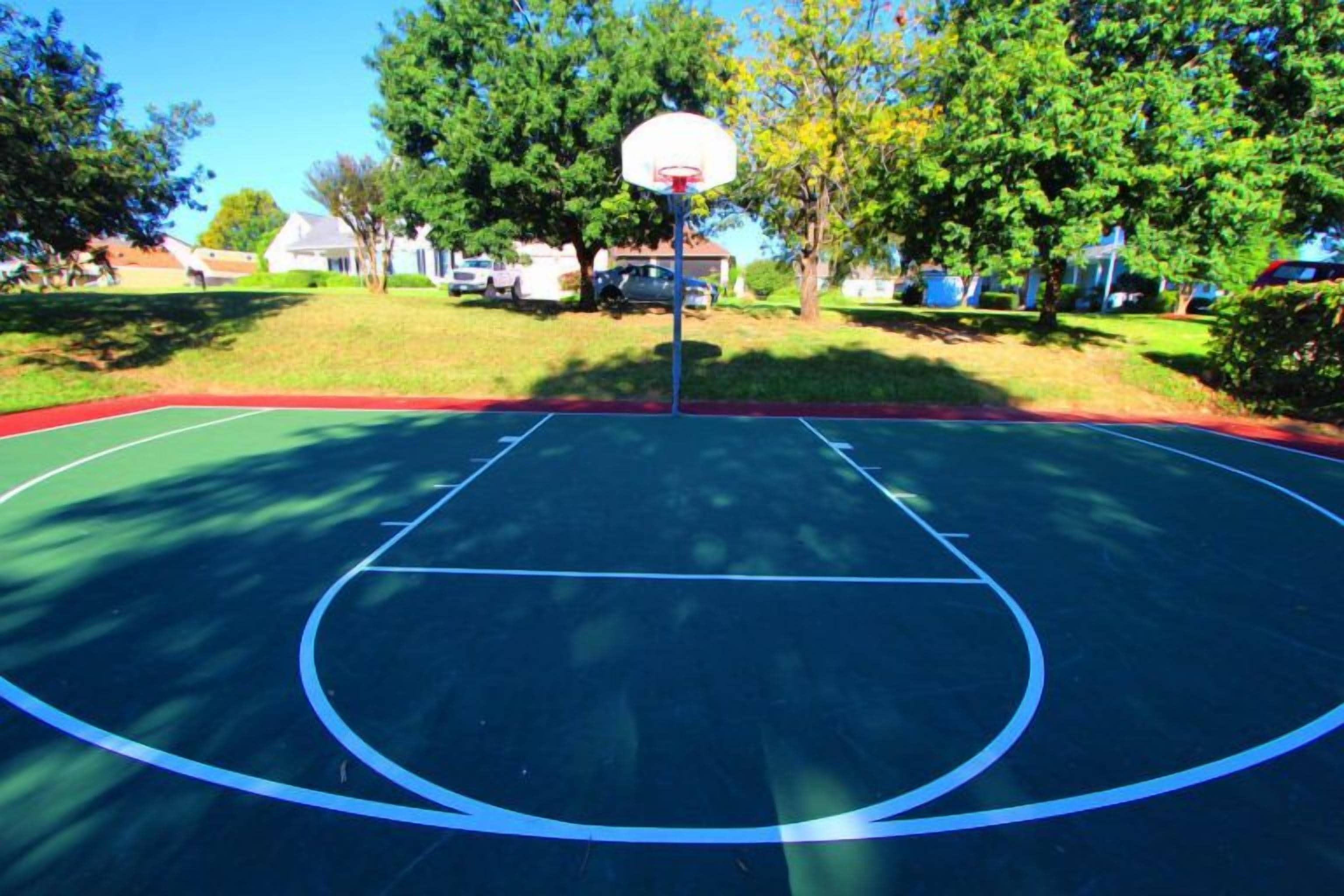An outdoor basketball court with a hoop, surrounded by trees and houses.