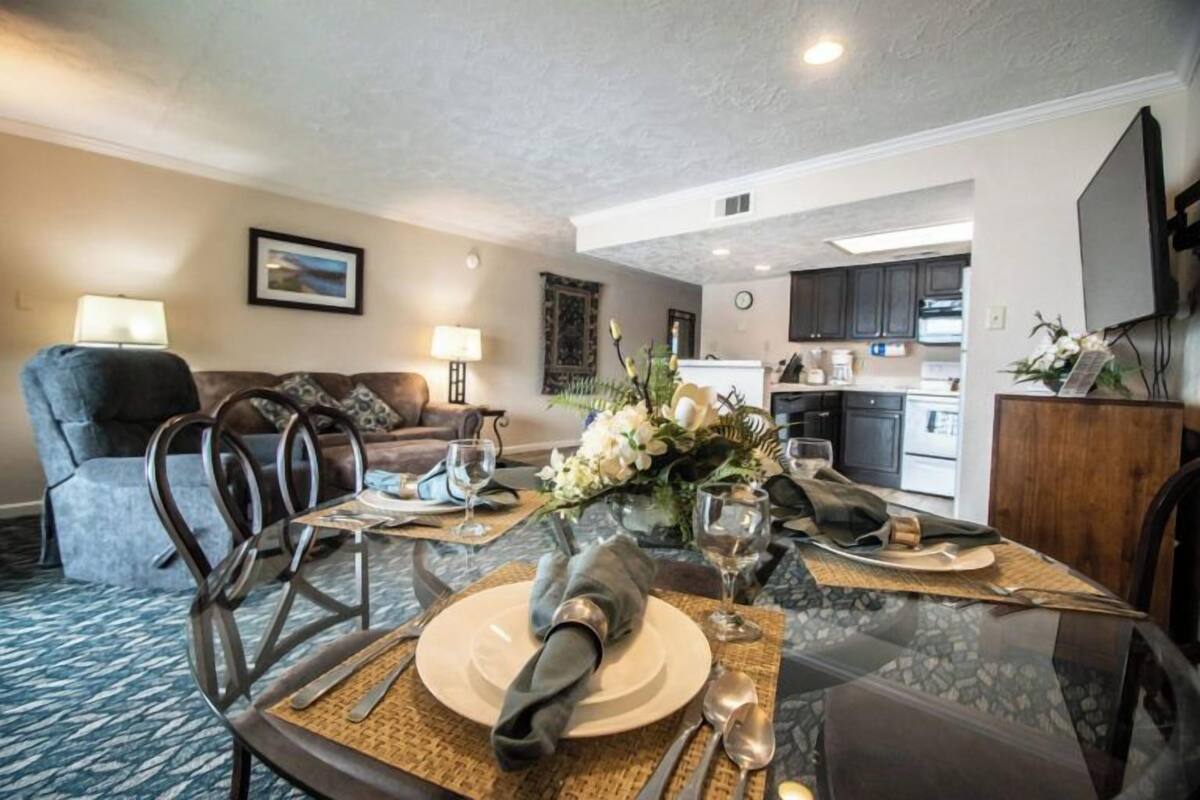 A dining area with a glass table set for four, a sofa, and a kitchen in the background.