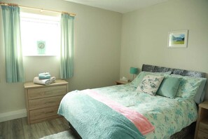 A bedroom with a bed, a nightstand, a dresser, a window with curtains, a wall clock, and a framed picture. - Killarney Town Center (Killarney)
