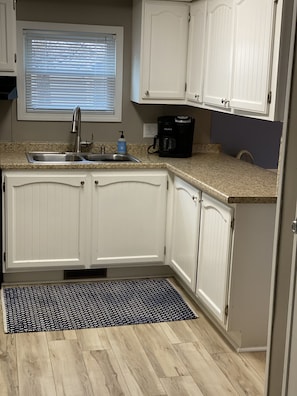 A kitchen with white cabinets, a granite countertop, a black coffee maker, and a window with blinds. - Just Peachy Peninsula Cottage -- Outside of Lakeside (LKSID MARBLHD)