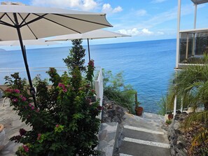 A view of the ocean from a balcony with a white umbrella, steps leading down to the water, and a stone wall.