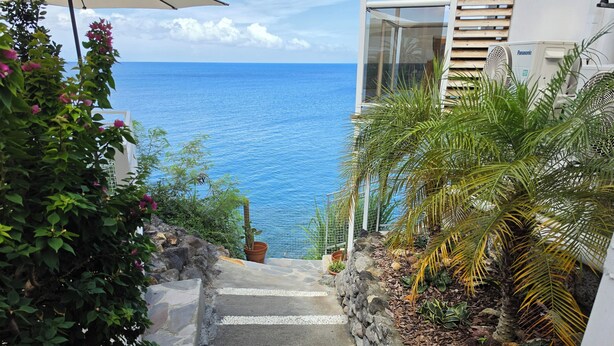 A staircase leading to a balcony overlooking the ocean, with a view of the sea and a white building with a Panasonic air conditioner. - Balisier Suite, sea and pool view. (Schoelcher)