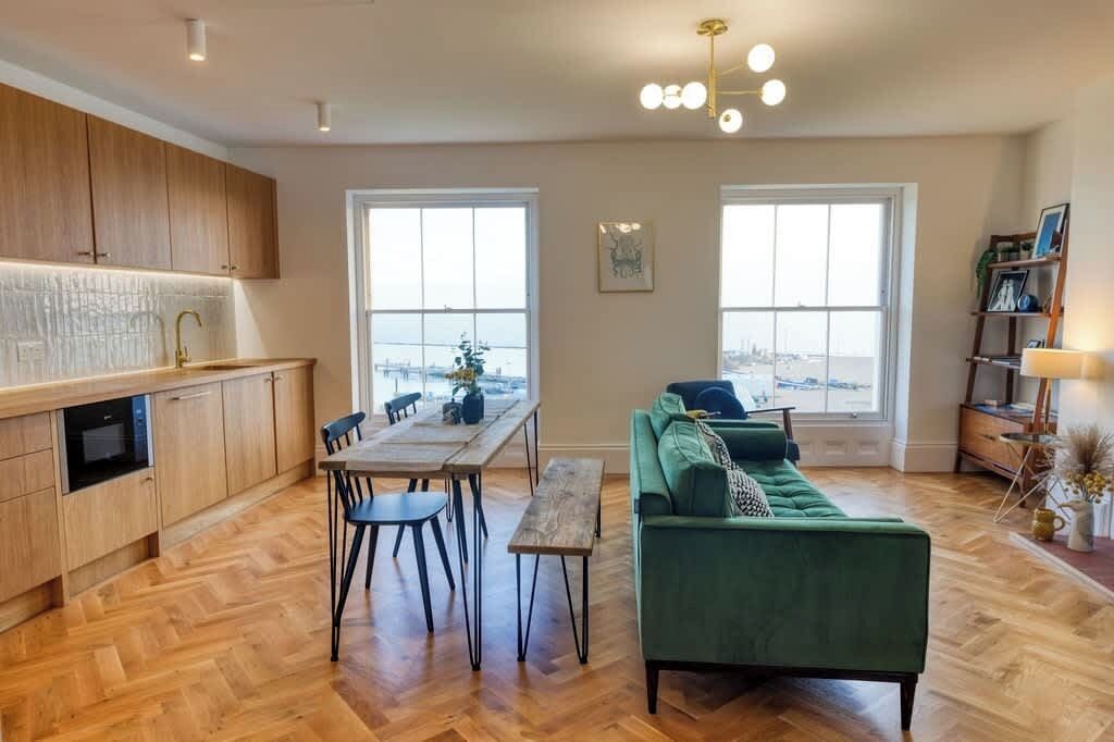 Une cuisine moderne avec des meubles en bois, une table à manger avec des chaises, un canapé vert et de grandes fenêtres offrant une vue dégagée.