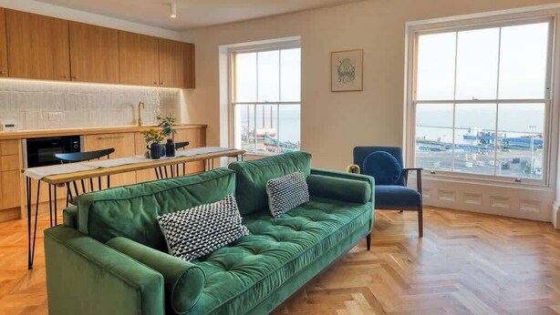 A modern kitchen with wooden cabinets, a dining area with a green sofa and a chair, and large windows with a view of the city. - The Lookout - Stylish penthouse with sea views (Ramsgate)