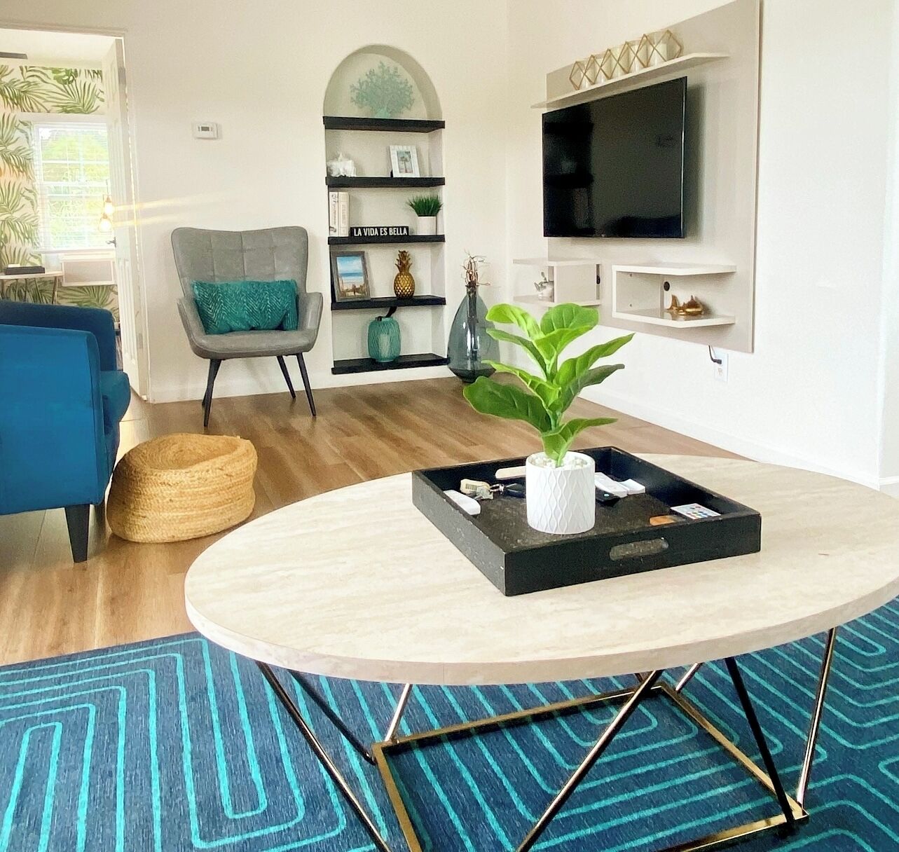 Una sala de estar moderna con una mesa redonda, un sofá azul, un sillón gris, una estantería y un televisor de pantalla plana montado en la pared.